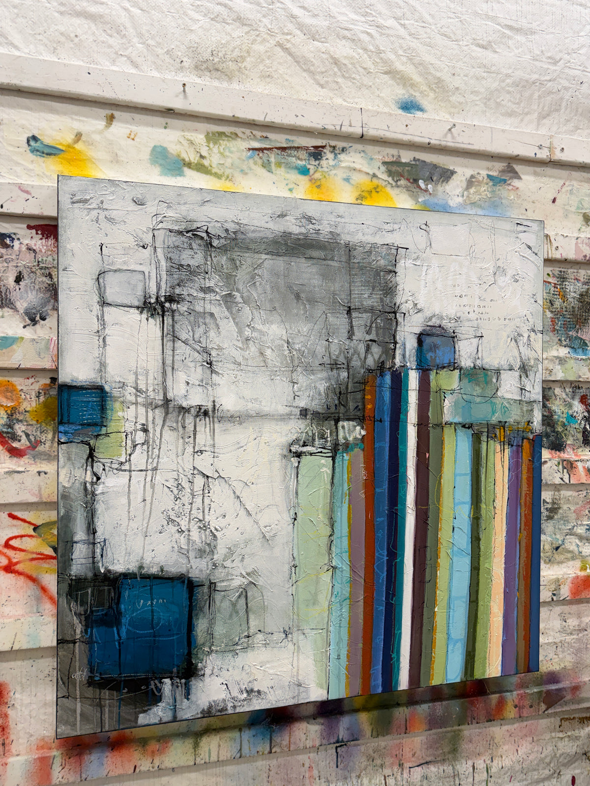 An abstract canvas painting featuring a textured gray and white background with vertical multicolored stripes in blue, green, yellow, orange, and red, displayed against a textured white wall with paint splatters.