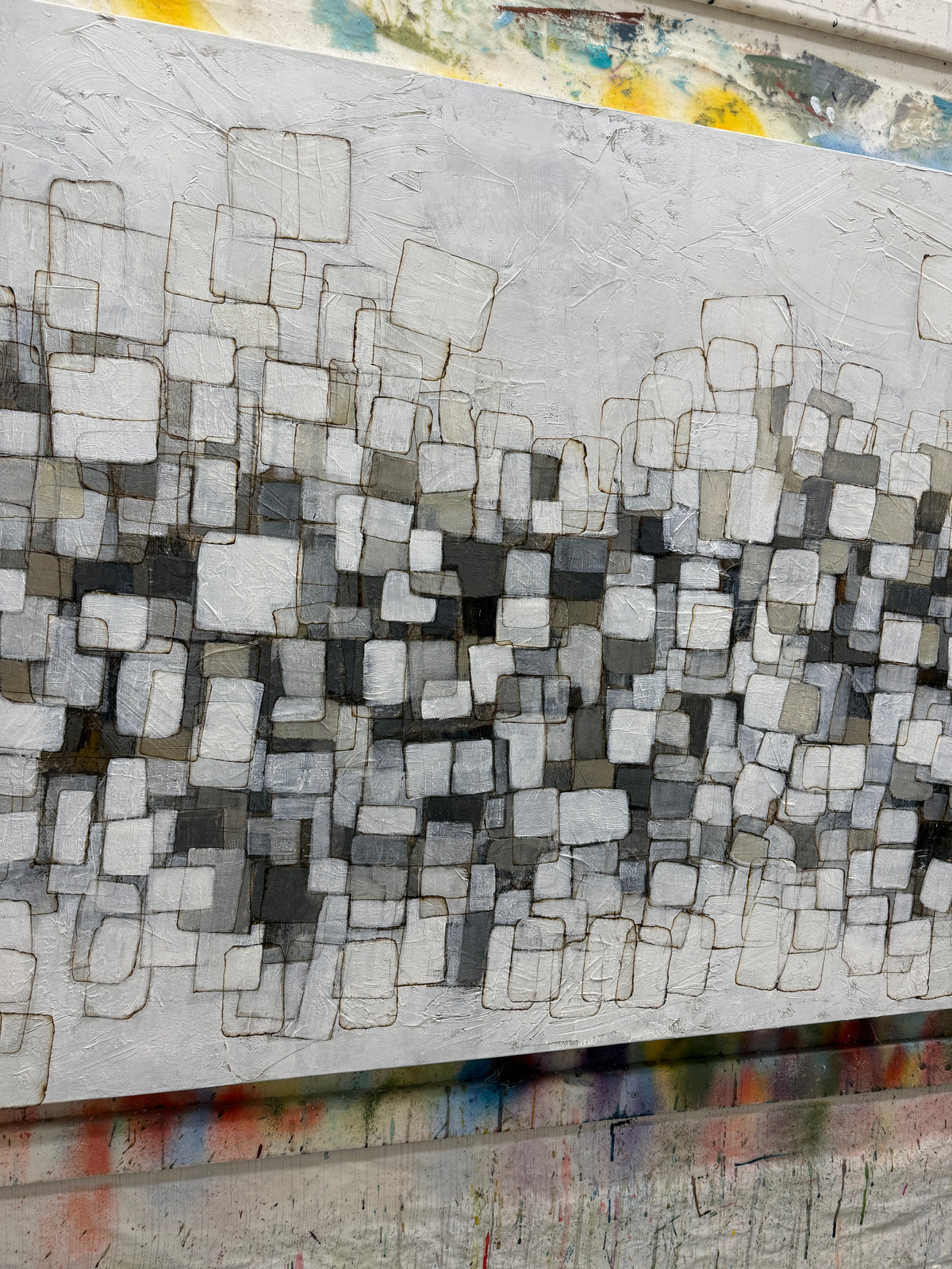 An abstract canvas painting featuring a textured grid of square and rectangular blocks in shades of gray, white, and charcoal, with visible paint drips and texture around the edges.