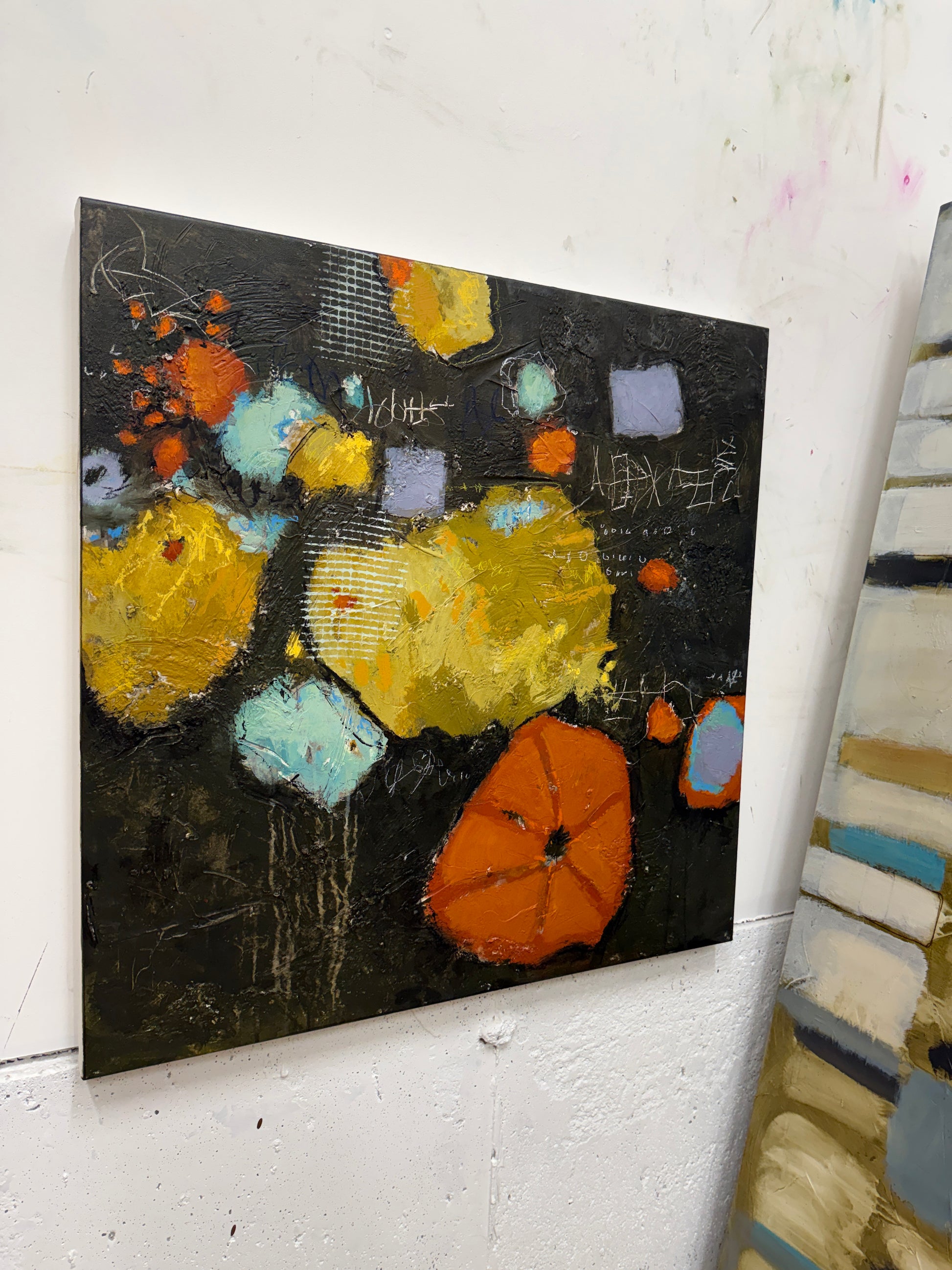 An abstract canvas painting featuring textured brushstrokes in yellow, orange, light blue, and lavender on a dark black background, hanging on a white wall.