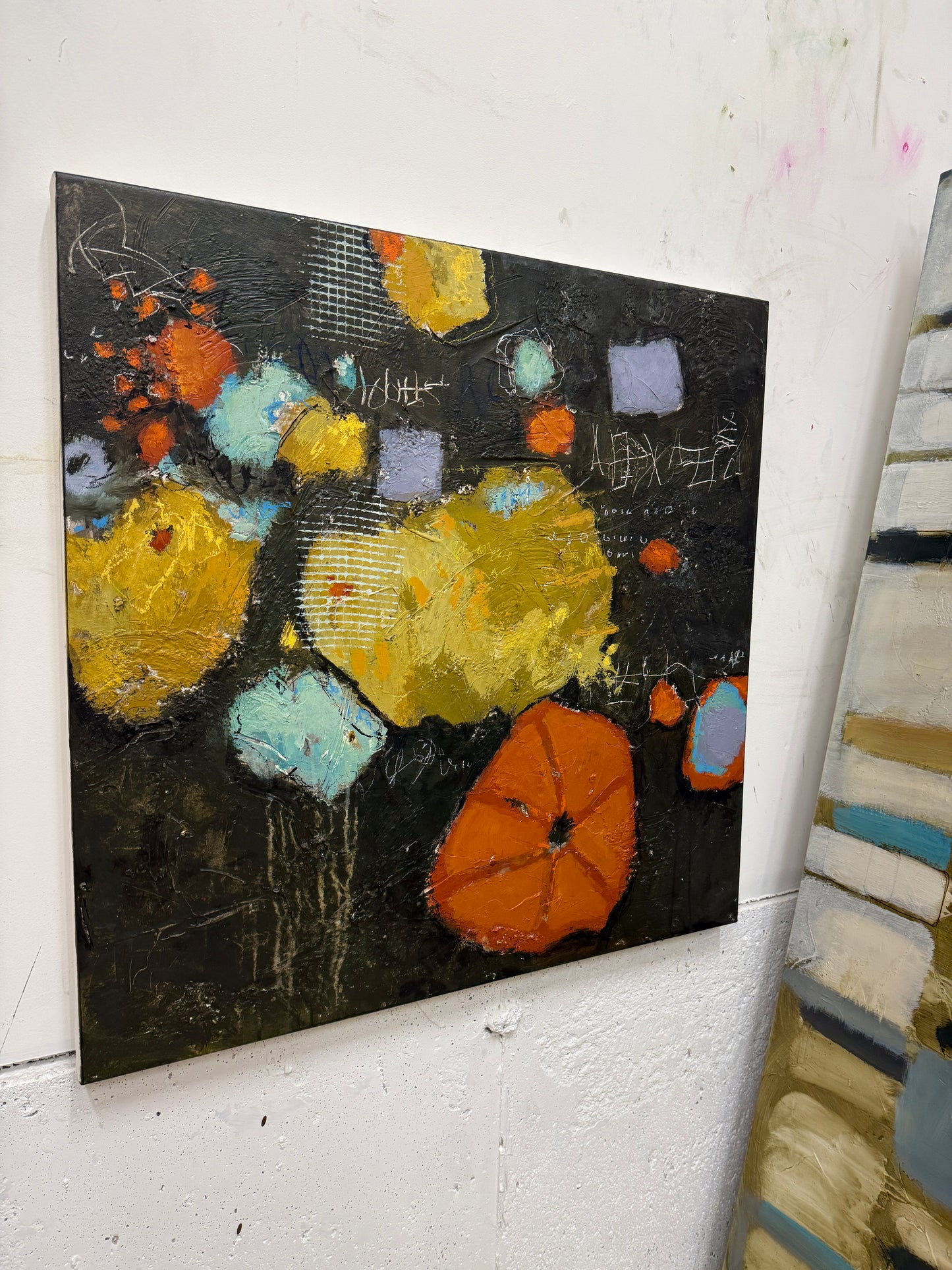 An abstract canvas painting featuring textured yellow, orange, and light blue shapes on a dark black background, hanging on a white wall next to other painted canvases.