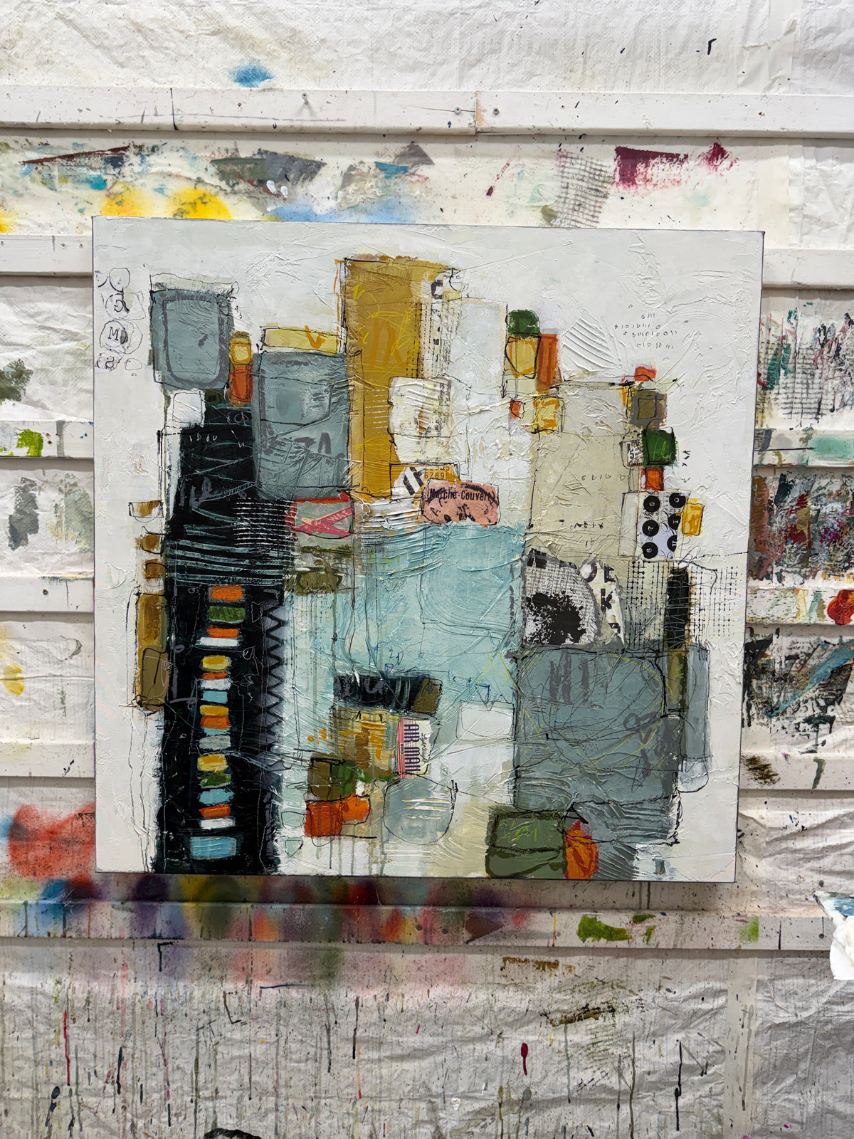 A 40 × 40-inch abstract mixed-media painting by Lori Mirabelli, featuring layered architectural shapes, textured white space, soft blue-grey blocks, mustard yellow, black, orange and green accents, with expressive linework and collage details.