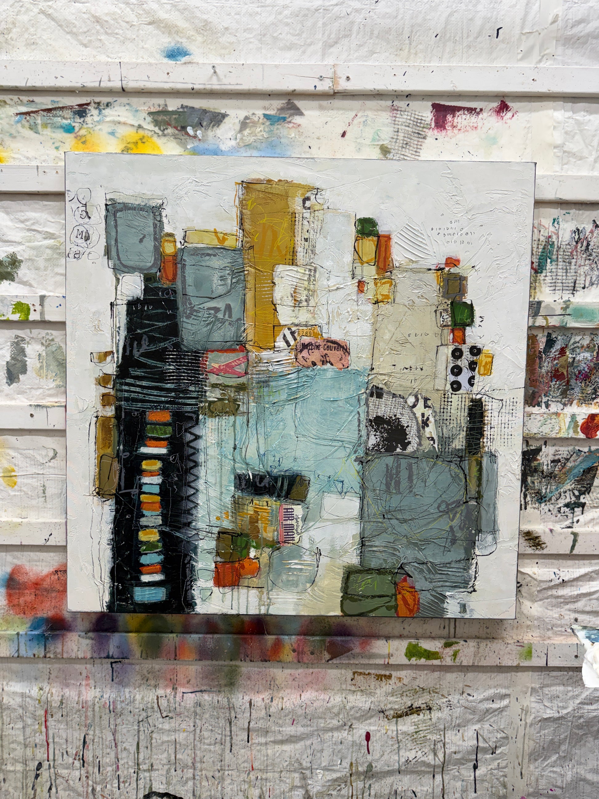 A 40 × 40-inch abstract mixed-media painting by Lori Mirabelli, featuring layered architectural shapes, textured white space, soft blue-grey blocks, mustard yellow, black, orange and green accents, with expressive linework and collage details.
