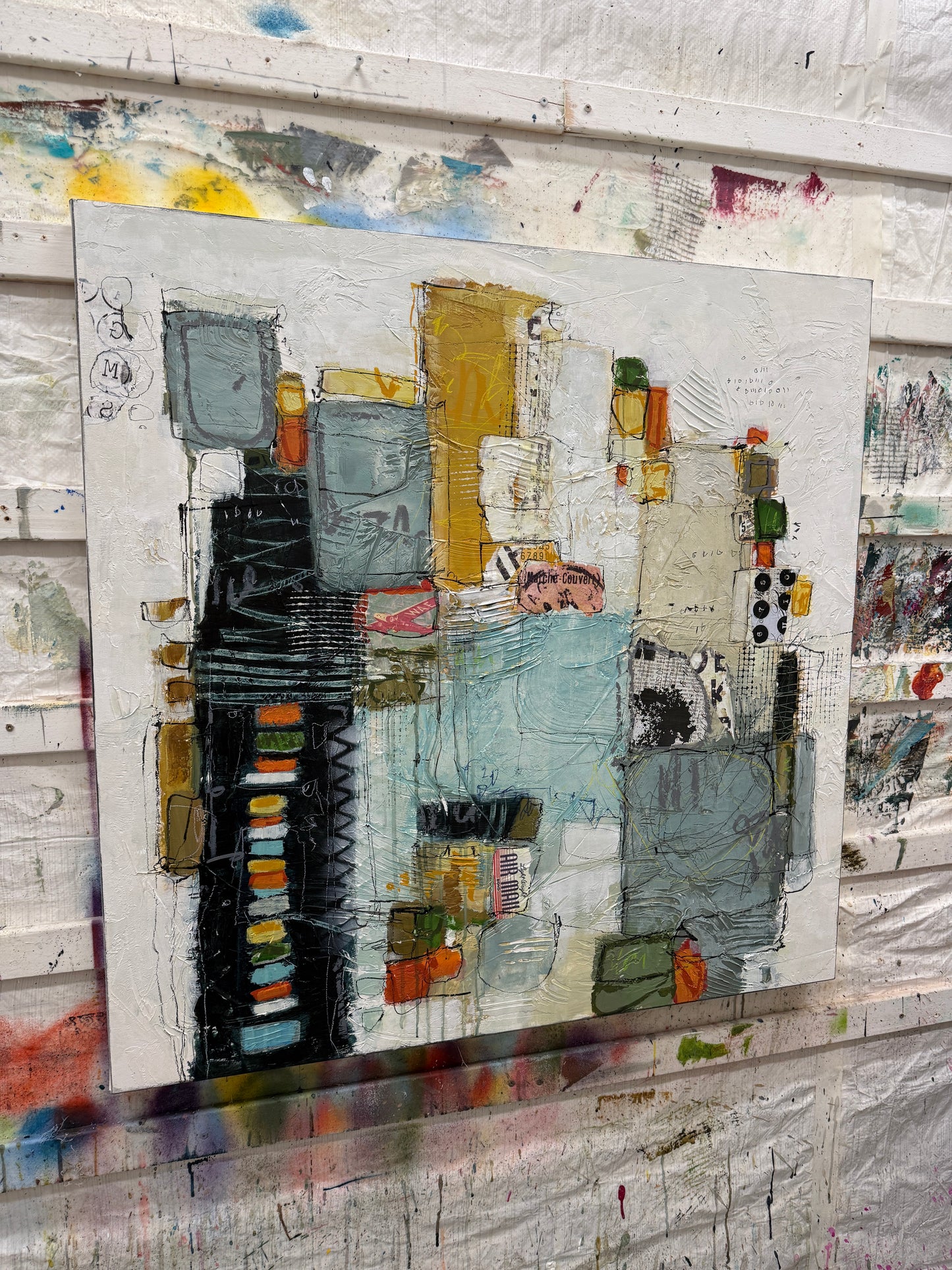 A 40 × 40-inch abstract mixed-media painting by Lori Mirabelli, featuring layered architectural shapes, textured white space, soft blue-grey blocks, mustard yellow, black, orange and green accents, with expressive linework and collage details.