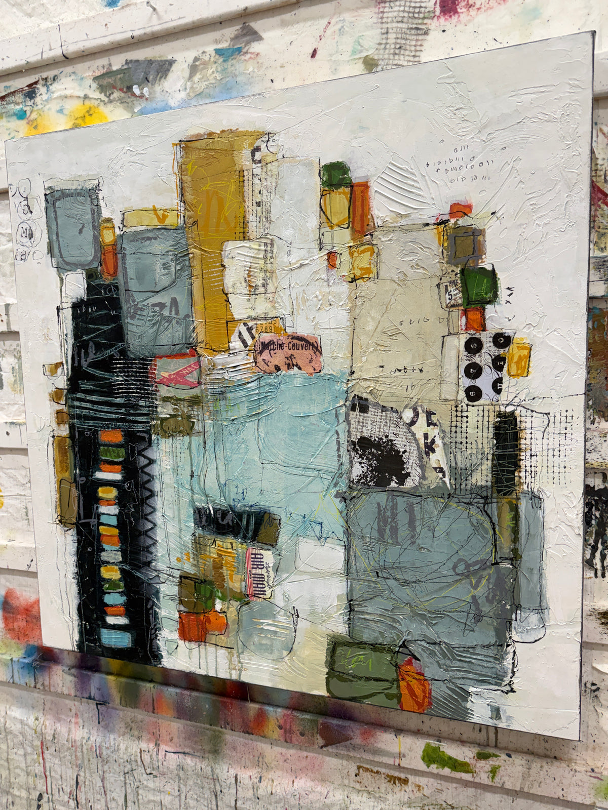 A 40 × 40-inch abstract mixed-media painting by Lori Mirabelli, featuring layered architectural shapes, textured white space, soft blue-grey blocks, mustard yellow, black, orange and green accents, with expressive linework and collage details.