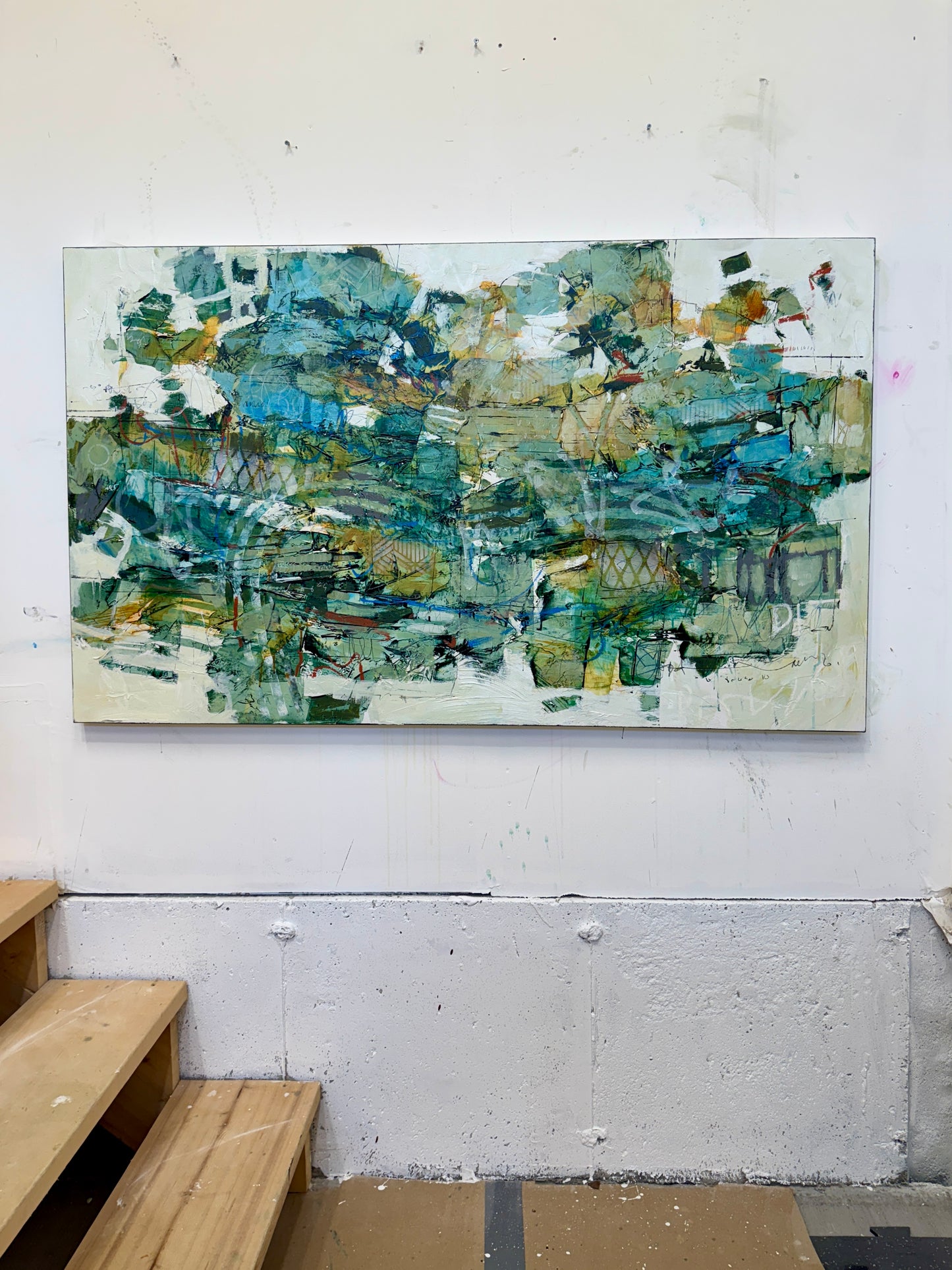 A large 36 x 60” abstract acrylic painting featuring layered greens, blues, whites, and hints of ochre. The piece has a dynamic, organic feel with visible mark-making and movement, blending structured lines with expressive brushwork. Displayed on a white studio wall above wooden steps. Lori mirabelli
