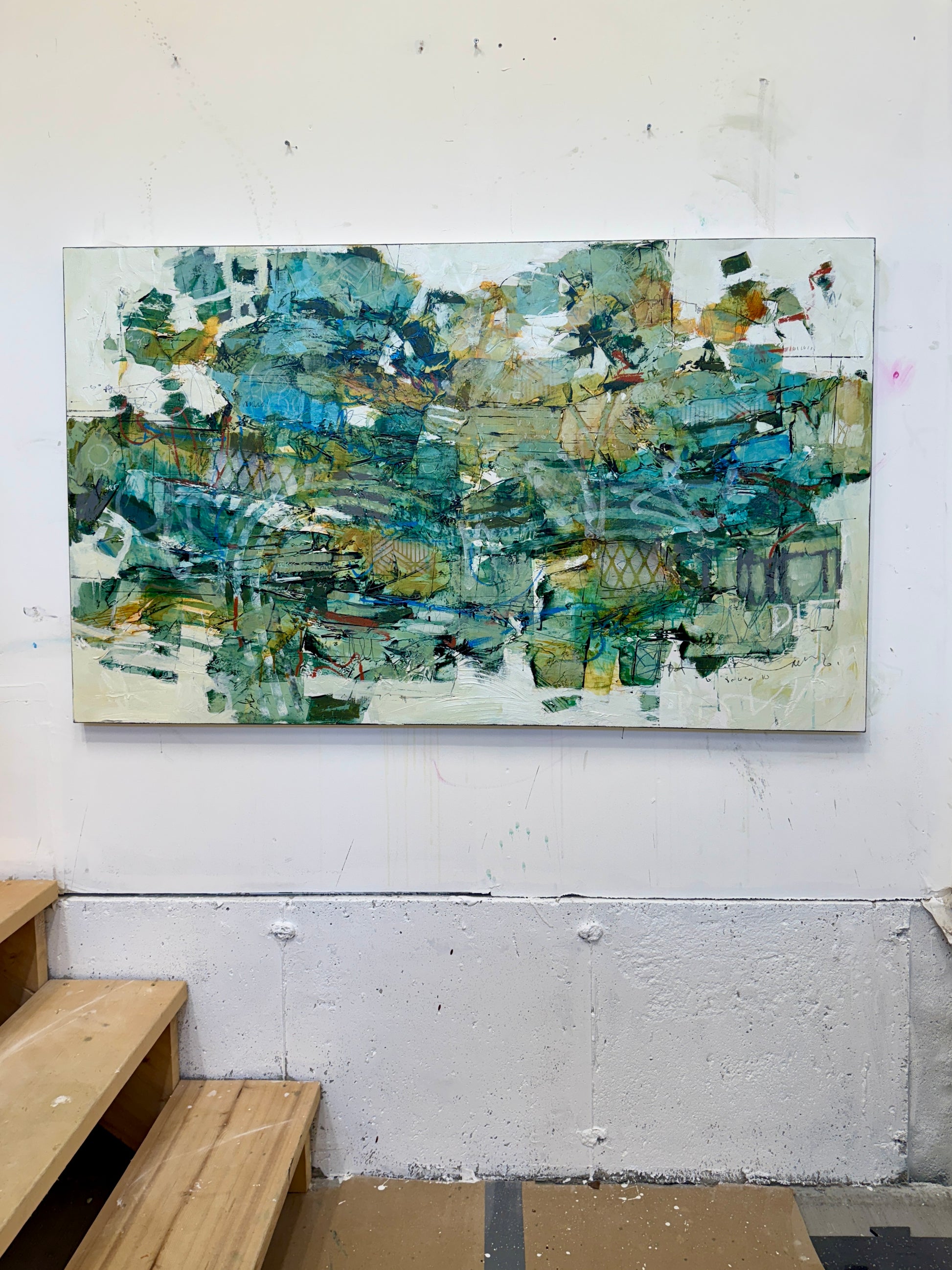 A large 36 x 60” abstract acrylic painting featuring layered greens, blues, whites, and hints of ochre. The piece has a dynamic, organic feel with visible mark-making and movement, blending structured lines with expressive brushwork. Displayed on a white studio wall above wooden steps. Lori mirabelli
