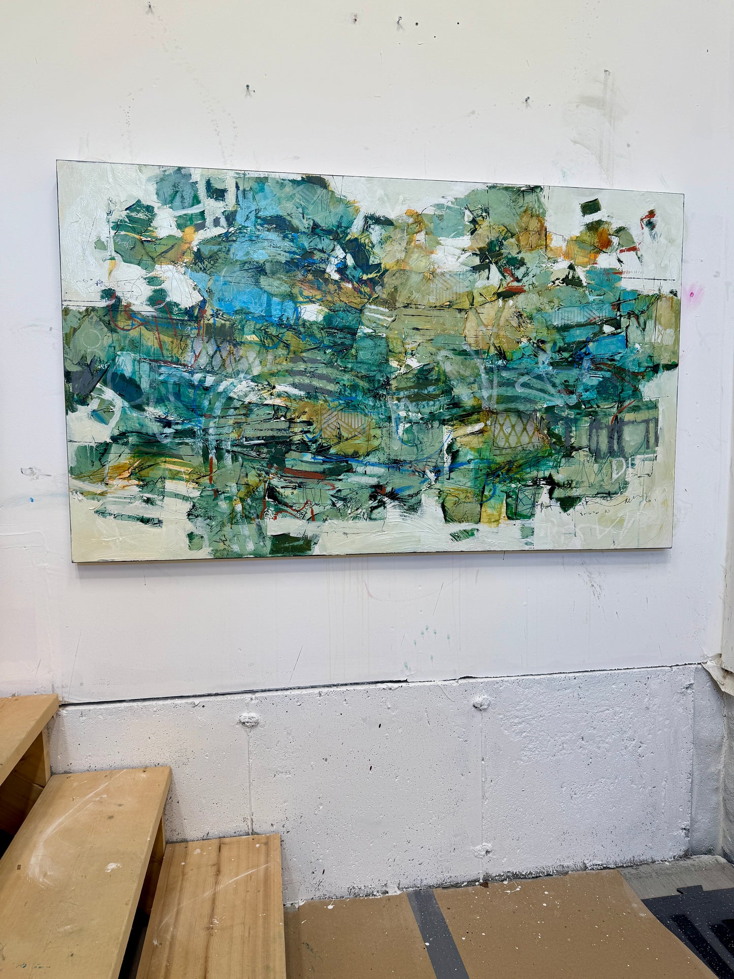 A large 36 x 60” abstract acrylic painting featuring layered greens, blues, whites, and hints of ochre. The piece has a dynamic, organic feel with visible mark-making and movement, blending structured lines with expressive brushwork. Displayed on a white studio wall above wooden steps. Lori mirabelli
