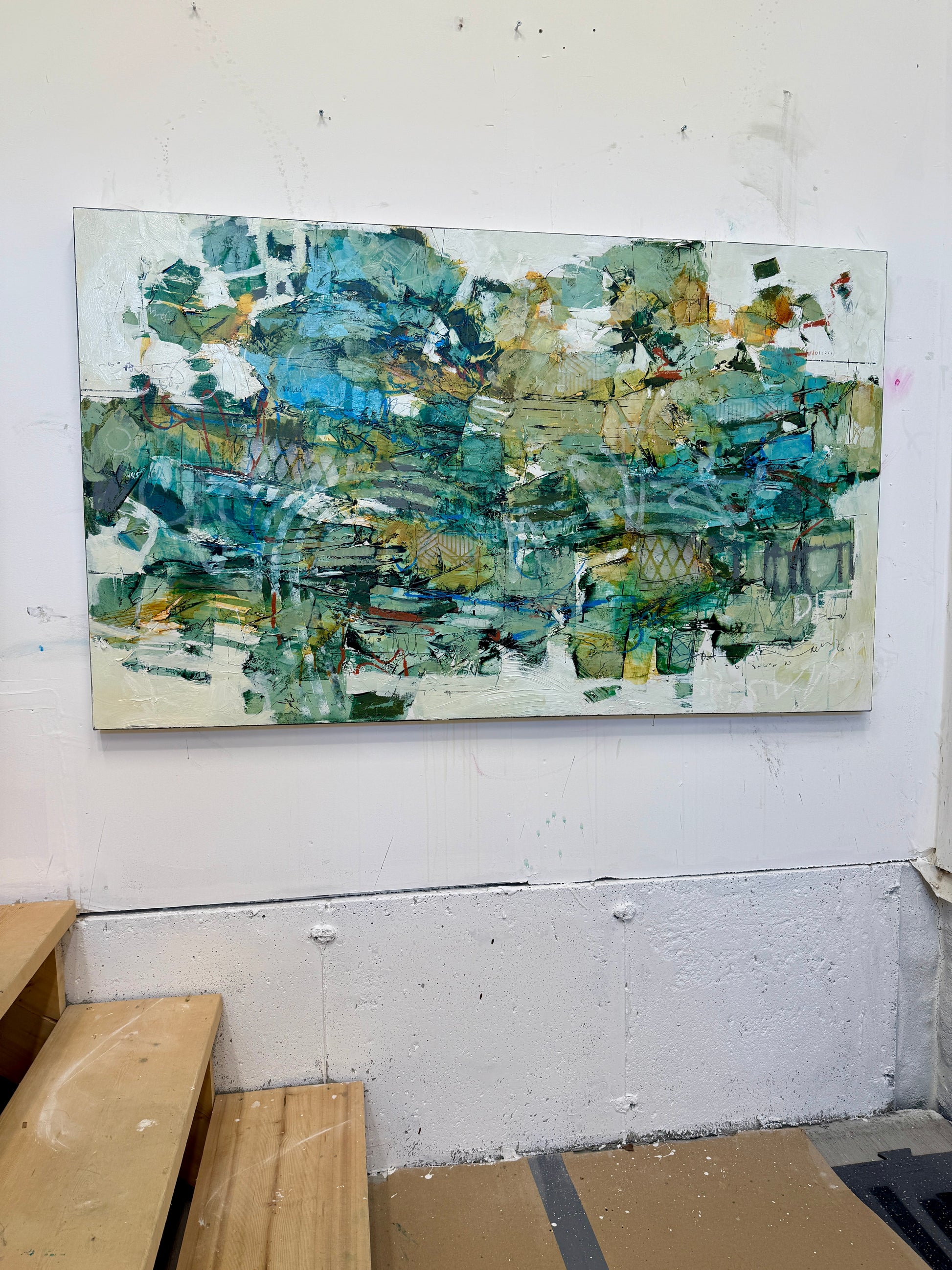 A large 36 x 60” abstract acrylic painting featuring layered greens, blues, whites, and hints of ochre. The piece has a dynamic, organic feel with visible mark-making and movement, blending structured lines with expressive brushwork. Displayed on a white studio wall above wooden steps. Lori mirabelli