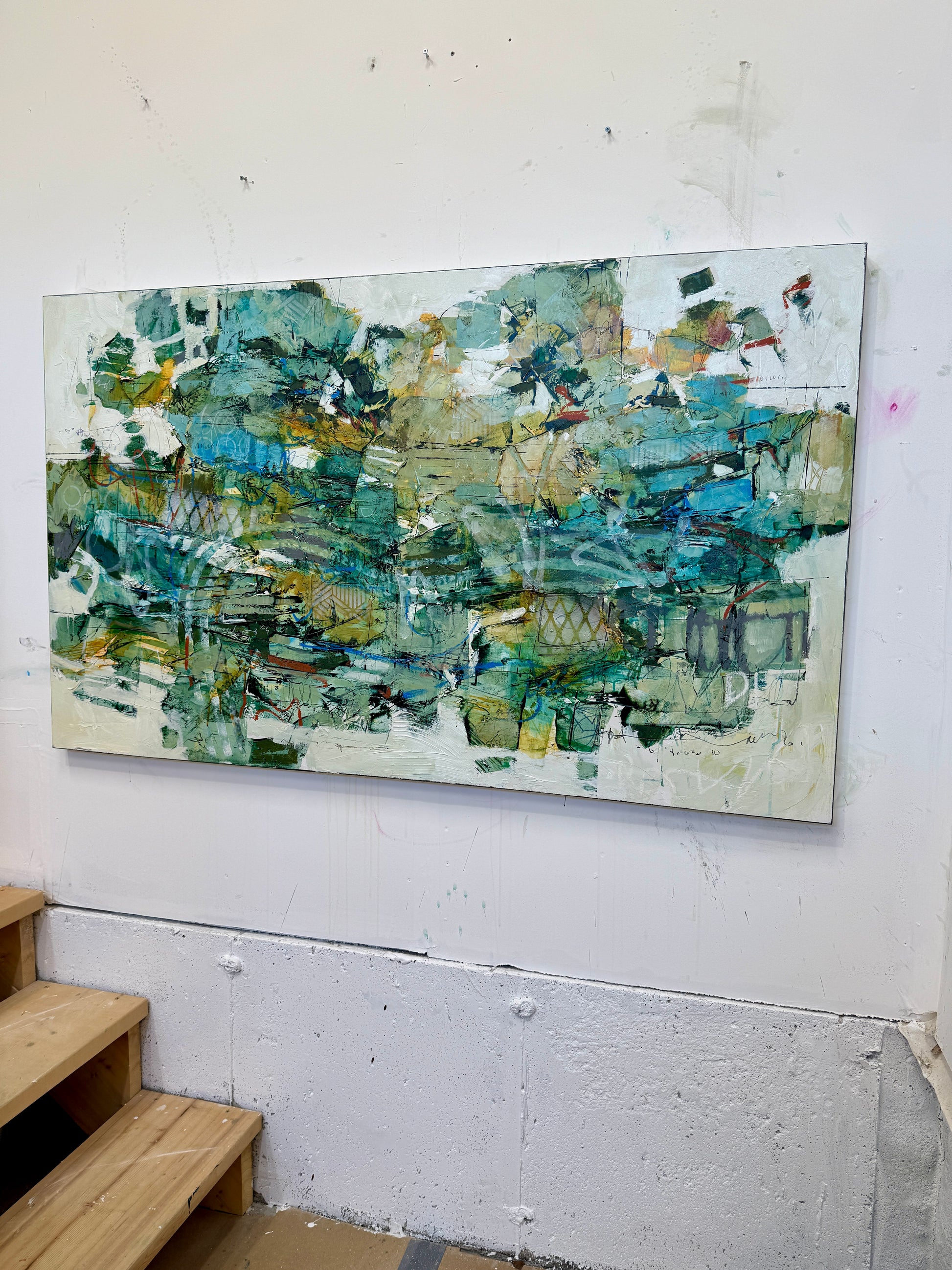 A large 36 x 60” abstract acrylic painting featuring layered greens, blues, whites, and hints of ochre. The piece has a dynamic, organic feel with visible mark-making and movement, blending structured lines with expressive brushwork. Displayed on a white studio wall above wooden steps. Lori mirabelli