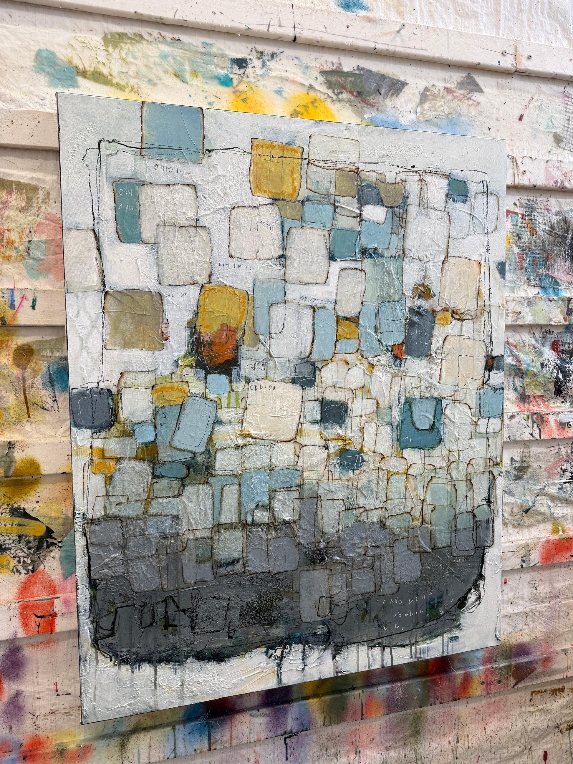 An abstract textured canvas painting with geometric blocks in shades of gray, blue, yellow, and white, displayed against a paint-splattered wall.