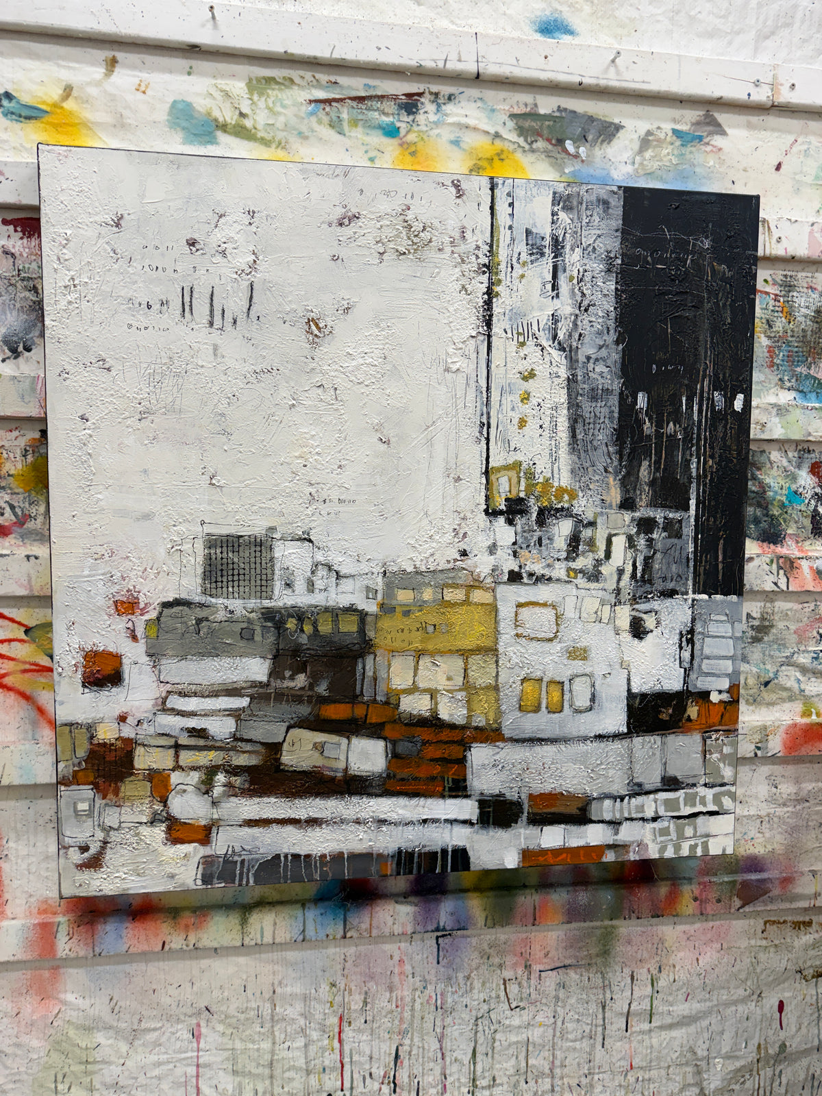 Square abstract painting with a large textured off-white area across the upper left. A charcoal-black section fills the upper right. Along the lower half, layered rectangular shapes and fine marks in grey, white, mustard yellow and rust orange suggest a loose cityscape.