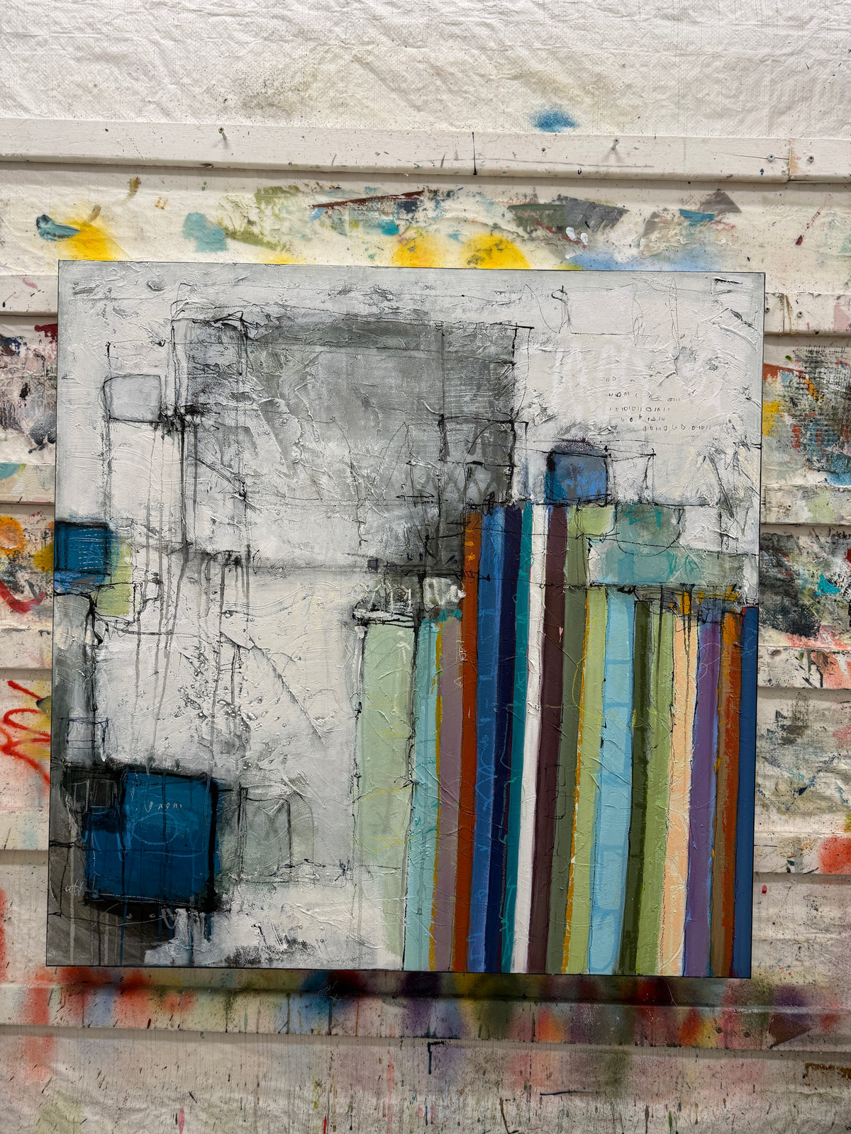 An abstract canvas painting featuring a textured gray and white background with vertical multicolored stripes in blue, green, yellow, and brown, displayed on a rustic, paint-splattered wooden wall.
