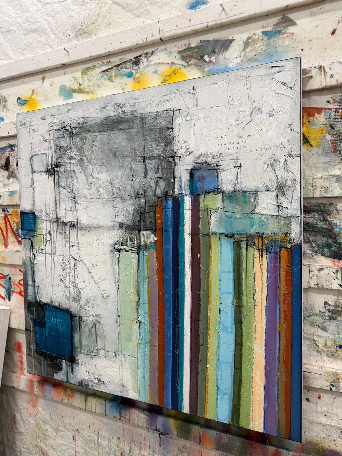 An abstract canvas painting with thick textured brushstrokes, featuring a white and gray background with vertical multicolored stripes in blue, green, orange, purple, and brown, displayed against a paint-splattered studio wall.