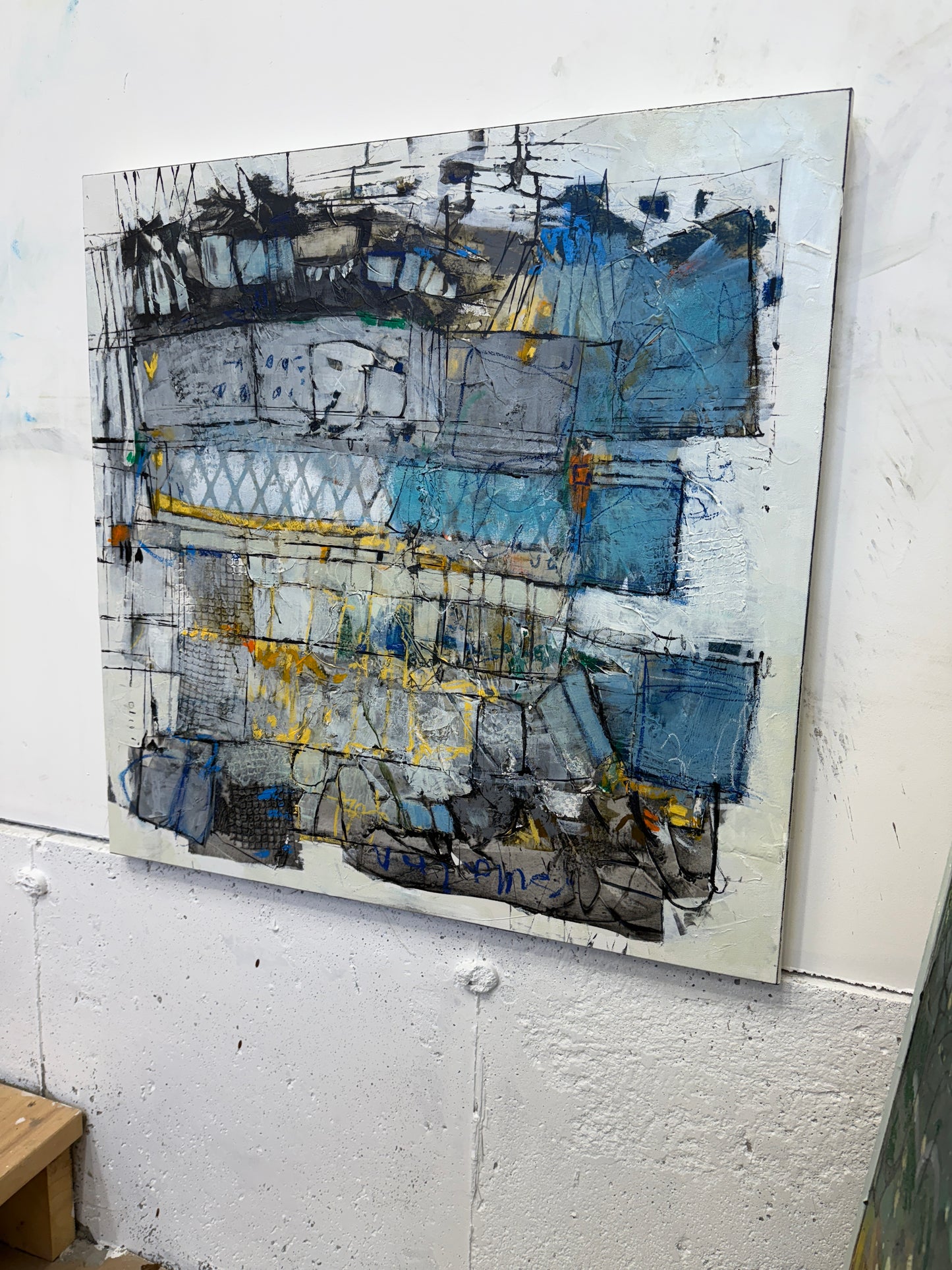 30x30 inch abstract acrylic painting on canvas featuring layered geometric shapes in grey, blue, and white with pops of bright yellow. The surface has rich texture, visible brushstrokes, and mark-making that gives it an urban, architectural feel with hints of movement and depth lori mirabelli