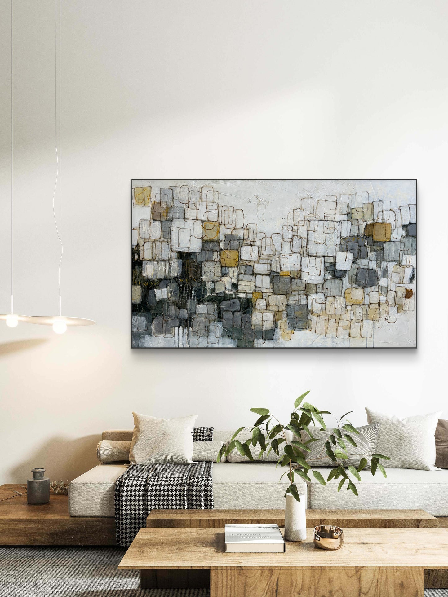 A framed abstract canvas print with a geometric block pattern in gray, white, black, and gold, displayed on a white wall above a modern beige sofa in a styled living room.