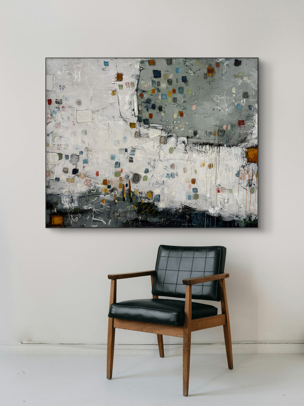 Large 48 × 60-inch abstract painting with textured white and sage-grey fields, scattered small squares in blue, mustard, rust, pink and green, and loose black and white lines. Dark charcoal layers anchor the bottom edge. The painting hangs above a black leather chair with wooden arms against a light wall.