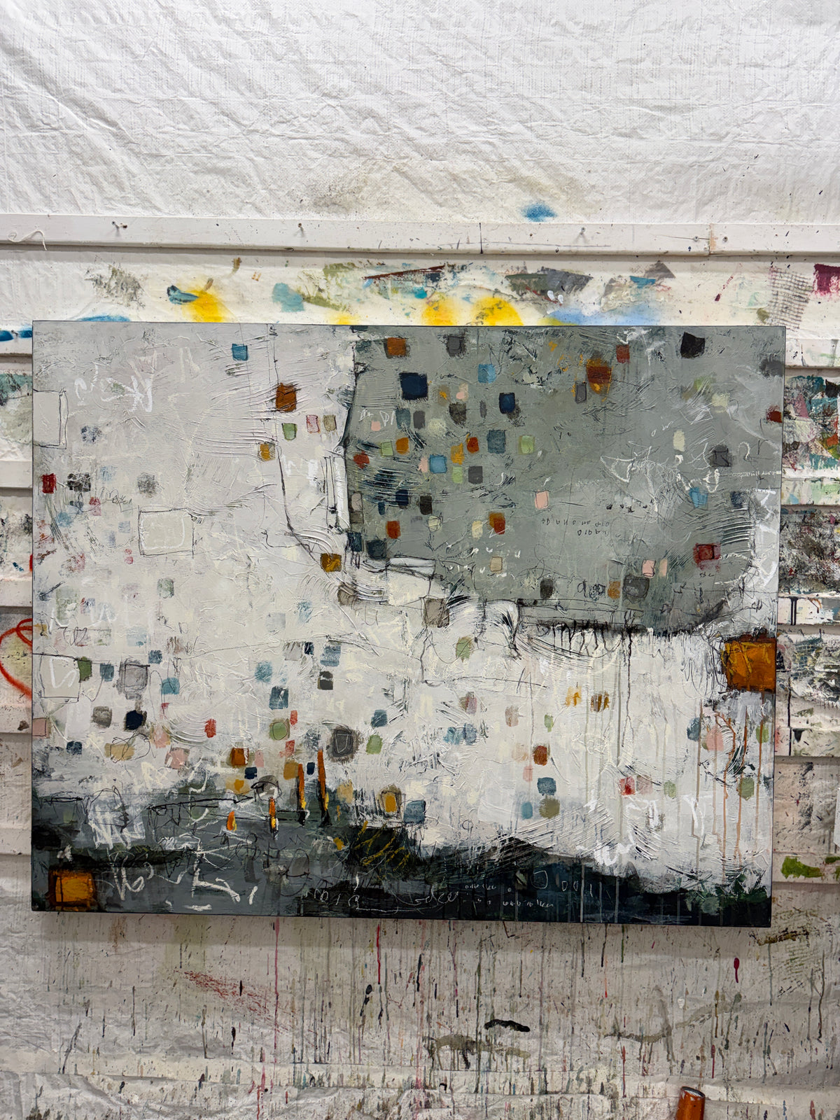 Large 48 × 60-inch abstract painting with textured white and sage-grey fields, scattered small squares in blue, mustard, rust, pink and green, and loose black and white lines. Dark charcoal layers anchor the bottom edge. The painting hangs above a black leather chair with wooden arms against a light wall.