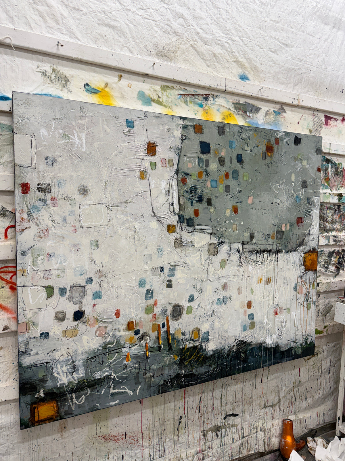 Large 48 × 60-inch abstract painting with textured white and sage-grey fields, scattered small squares in blue, mustard, rust, pink and green, and loose black and white lines. Dark charcoal layers anchor the bottom edge. The painting hangs above a black leather chair with wooden arms against a light wall.