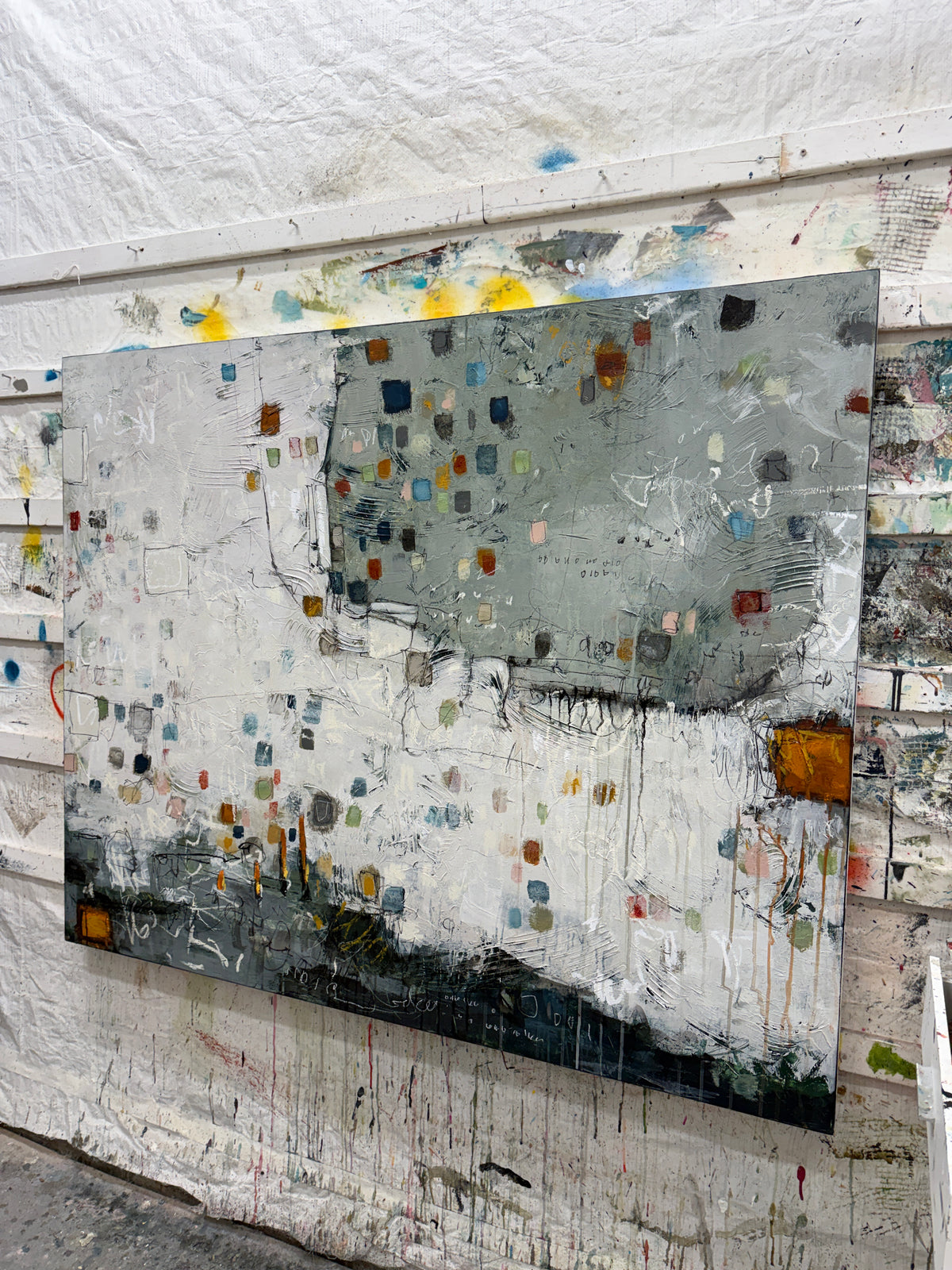 Large 48 × 60-inch abstract painting with textured white and sage-grey fields, scattered small squares in blue, mustard, rust, pink and green, and loose black and white lines. Dark charcoal layers anchor the bottom edge. The painting hangs above a black leather chair with wooden arms against a light wall.
