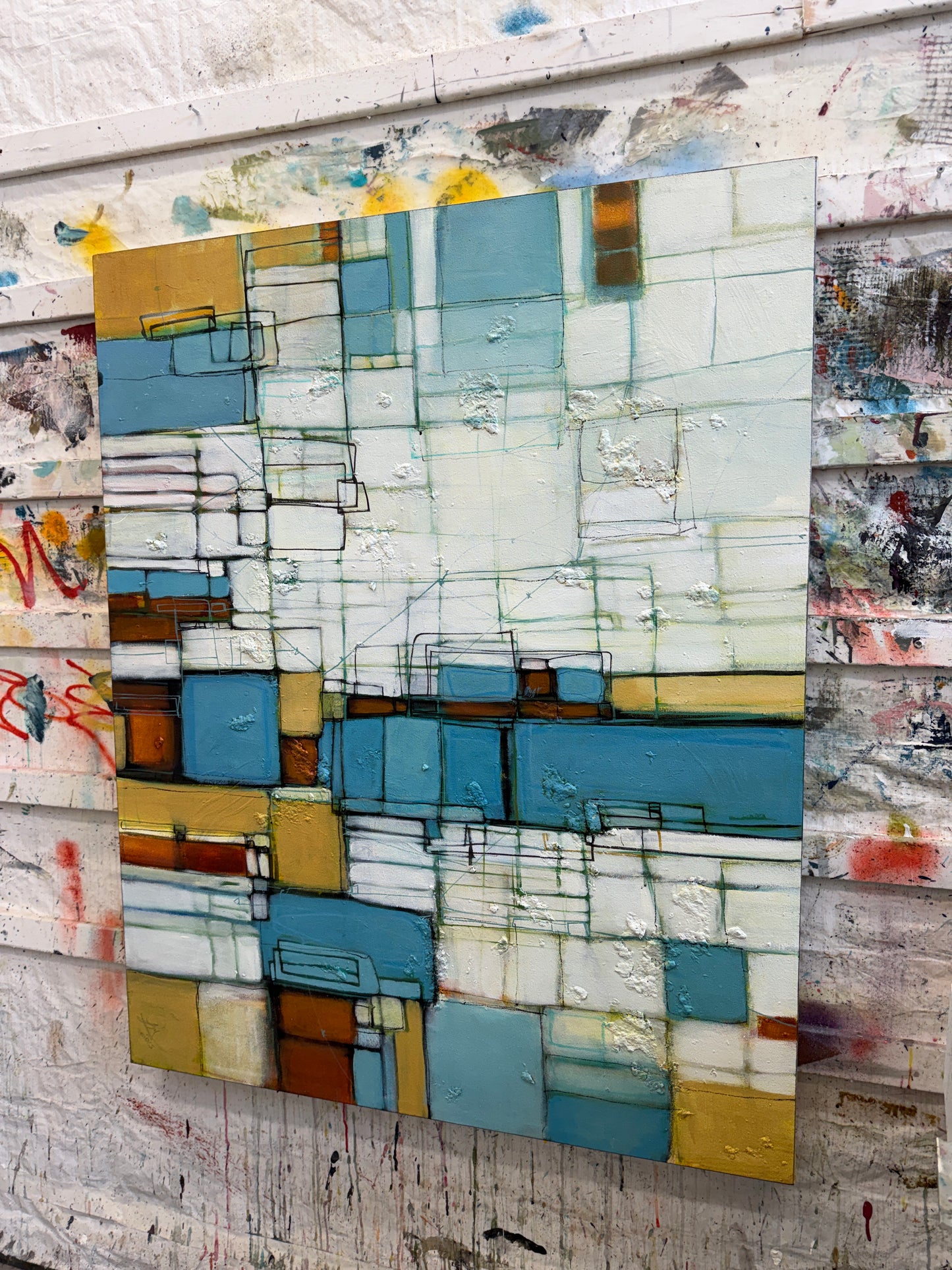 An abstract geometric canvas painting with blocks of blue, beige, white, and brown, displayed against a rustic, paint-splattered wooden background.