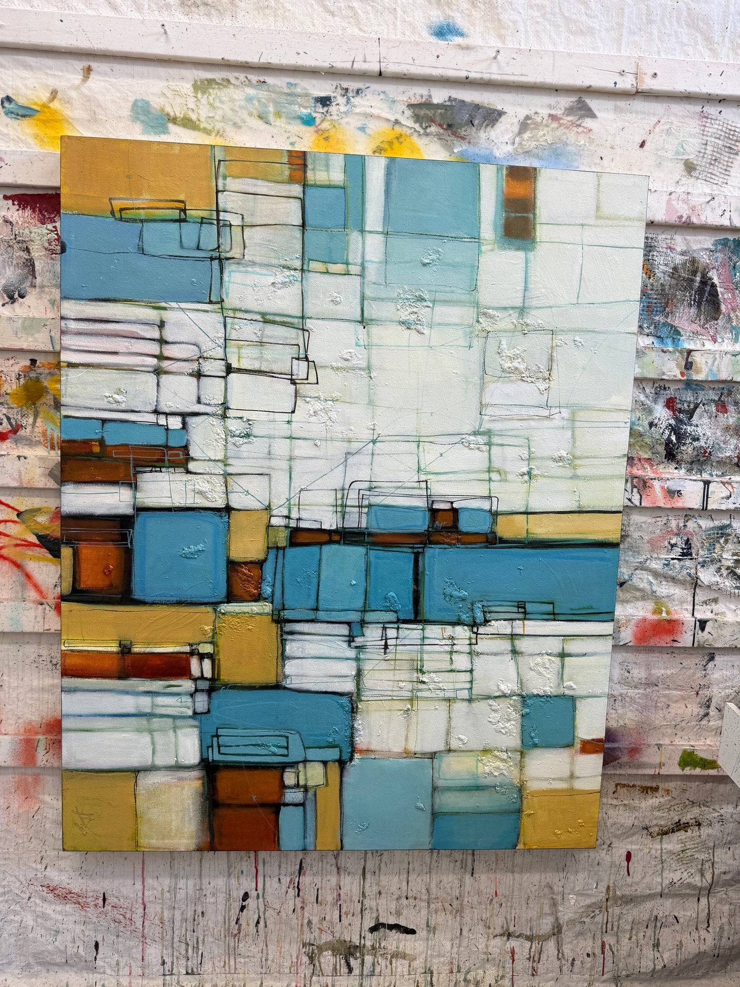An abstract geometric canvas painting with blocks of light blue, teal, white, mustard yellow, and brown, displayed against a textured, paint-splattered white wall.