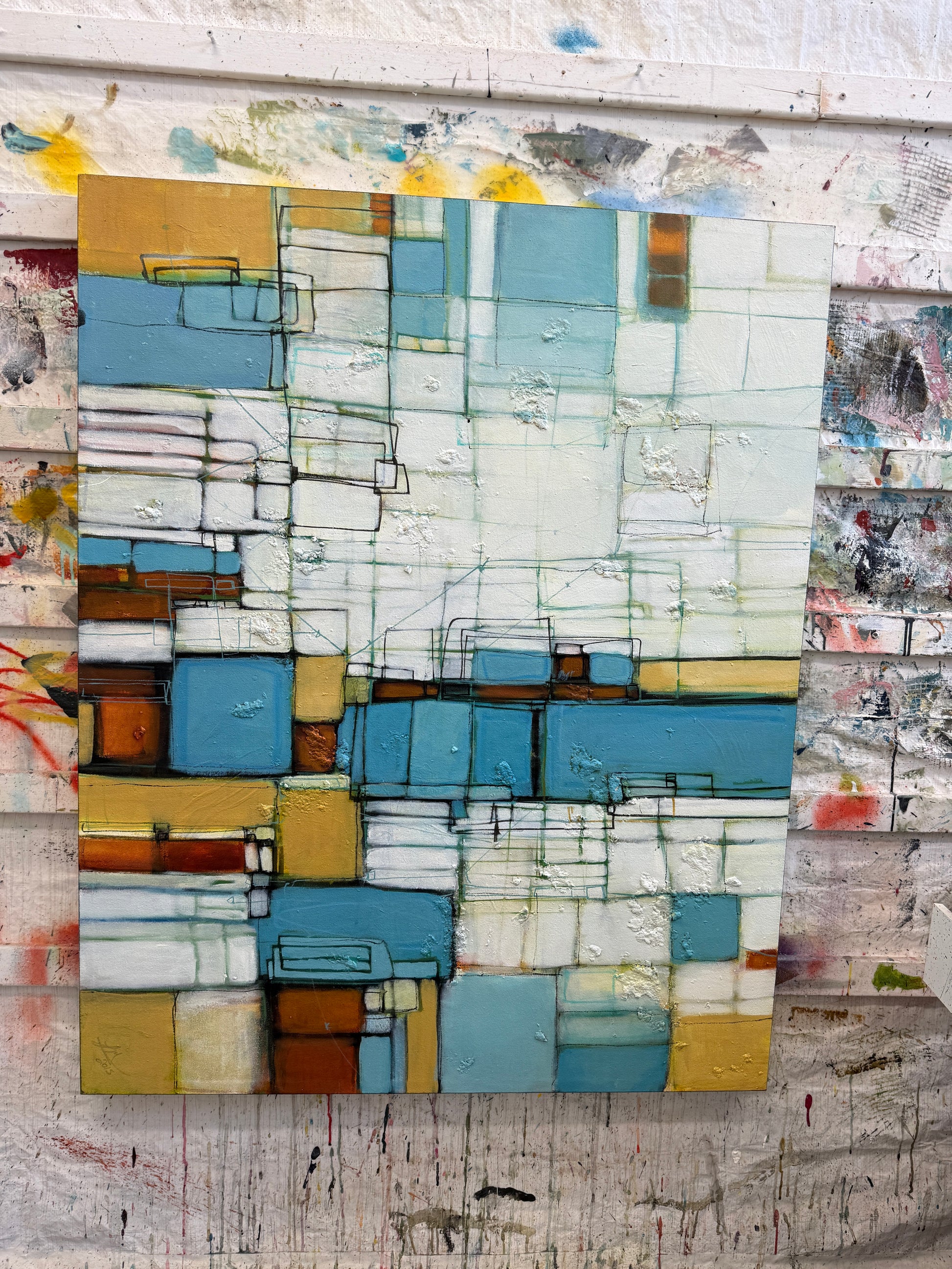 An abstract geometric canvas painting with blocks of light blue, teal, white, mustard yellow, and brown, displayed against a textured, paint-splattered white wall.