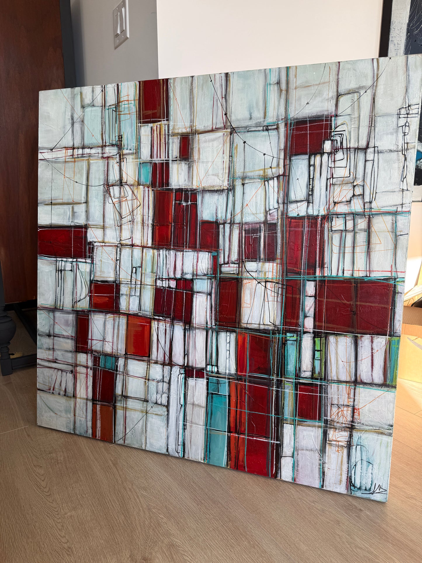 An abstract geometric canvas painting with a grid pattern of red, white, and teal blocks, leaning against a wall on a light wood floor.