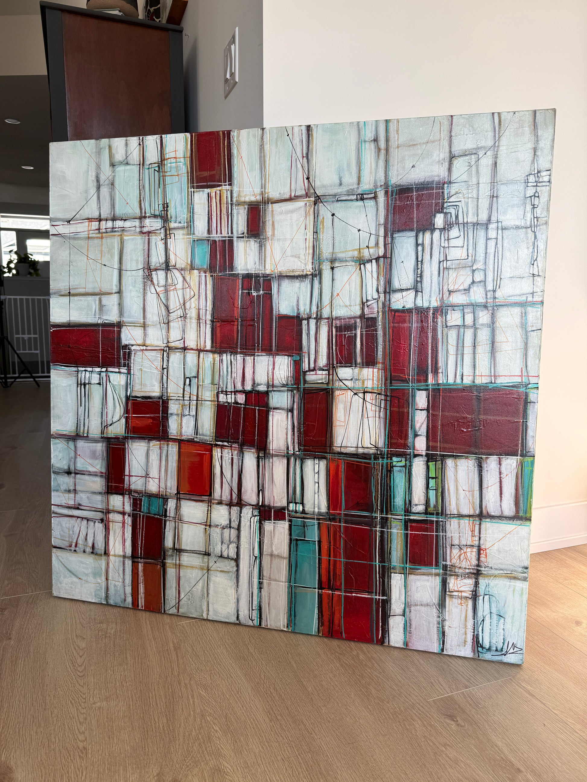 A large abstract canvas painting with a geometric block pattern in red, white, gray, and teal, leaning against a wall on a light wood floor.