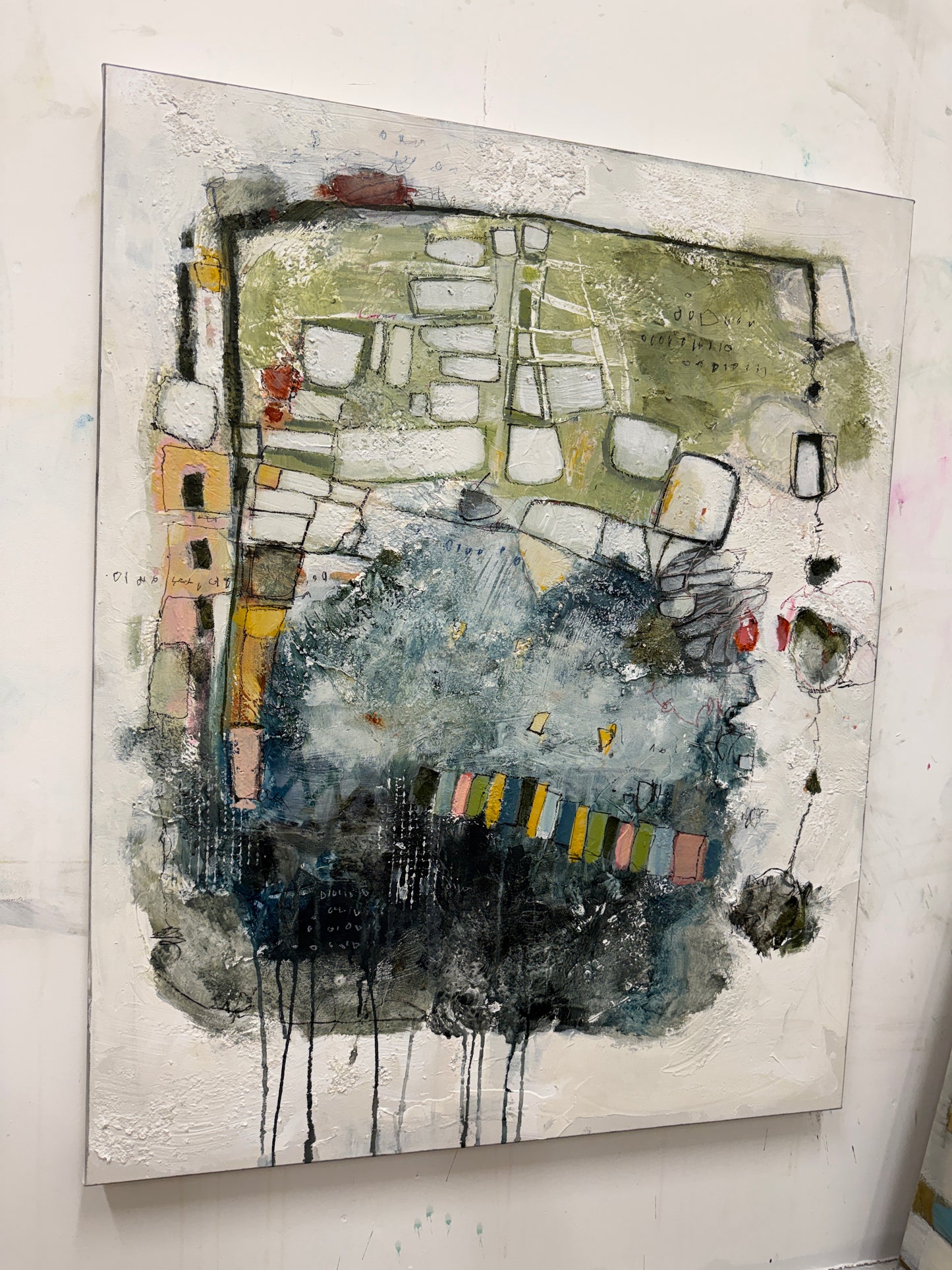 An abstract mixed-media painting on canvas, featuring textured splashes of blue, green, black, white, and yellow, displayed against a white wall.