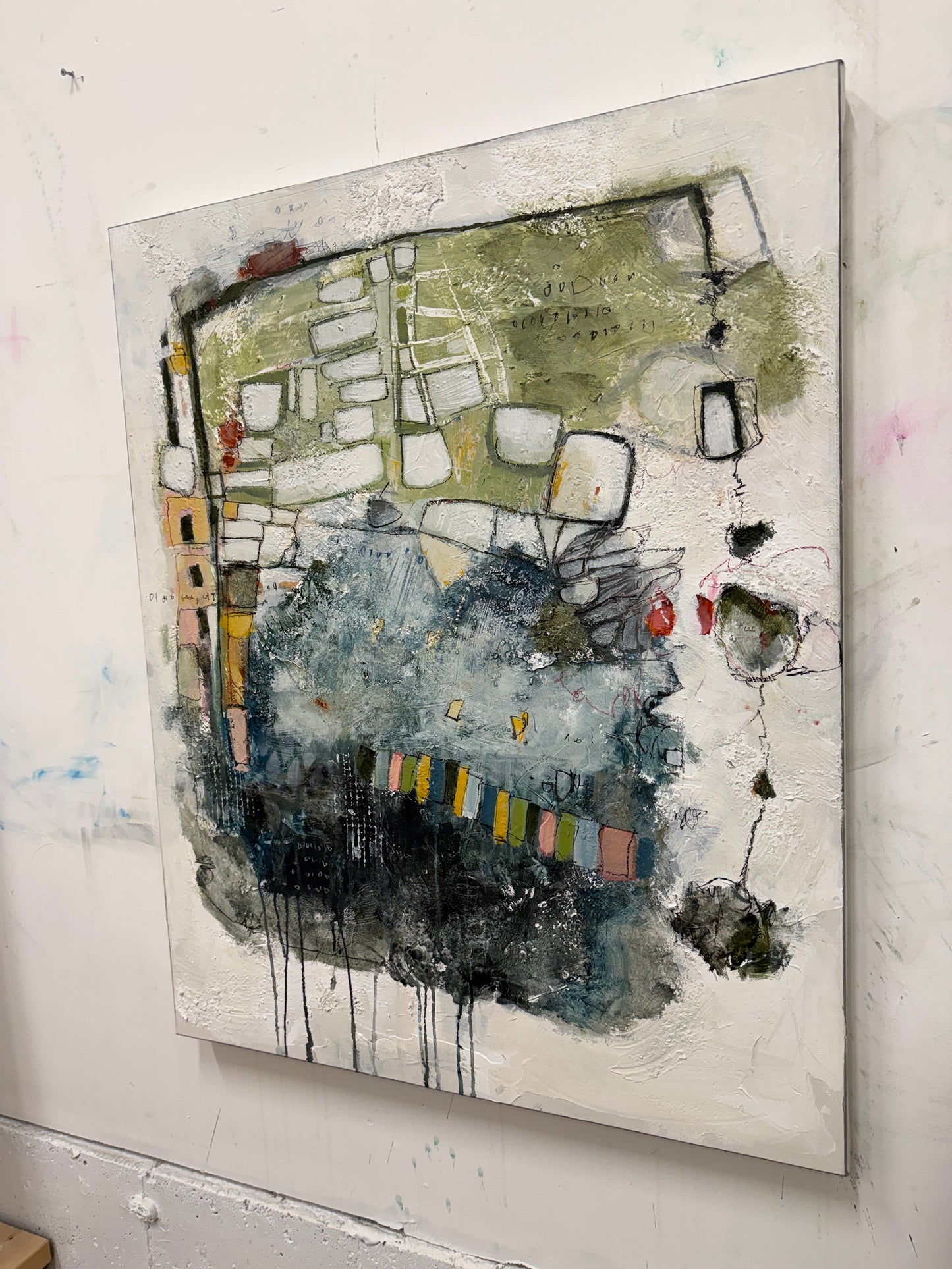 An abstract mixed-media painting on a square canvas, featuring textured layers of olive green, slate blue, charcoal black, and white with small accents of yellow, red, and orange, displayed on a white wall.