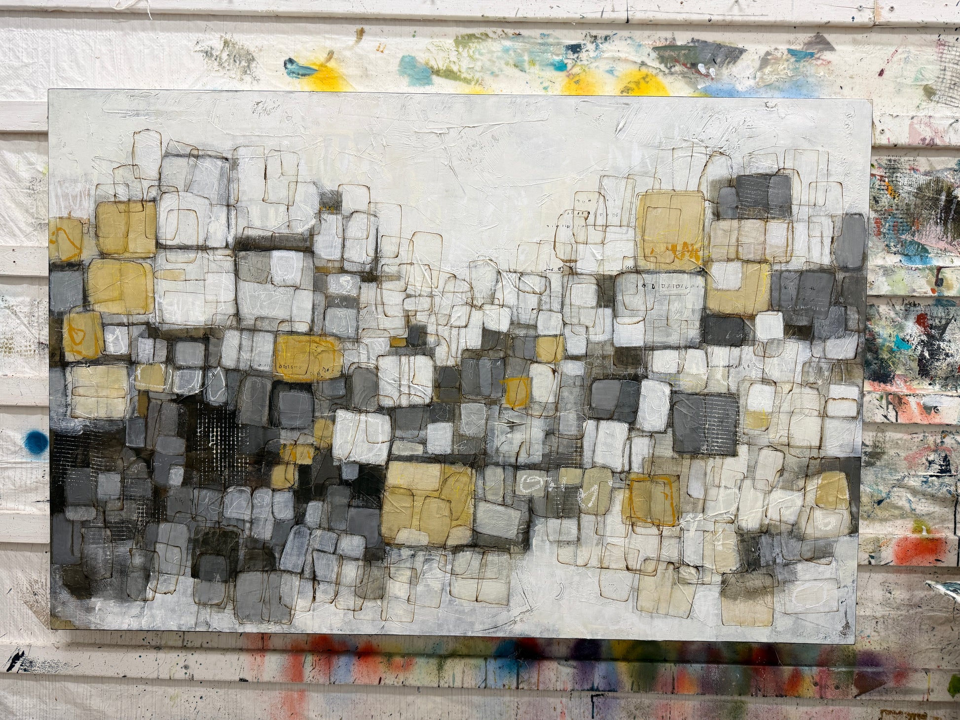 An abstract mixed-media painting featuring a grid-like pattern of overlapping rectangles in shades of gray, white, black, and muted yellow, displayed on a rustic, paint-splattered wooden wall.