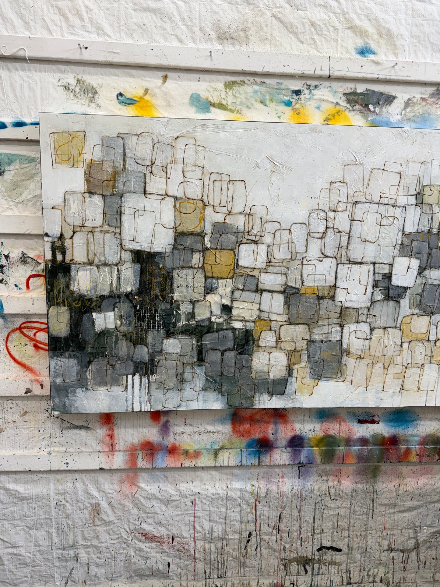 An abstract canvas painting featuring a textured grid of white, gray, black, and muted gold blocks, displayed against a white wall with colorful paint splatters and drips.